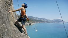 A woman climbs a rock wall with a pretty bay and sailboats in the background.