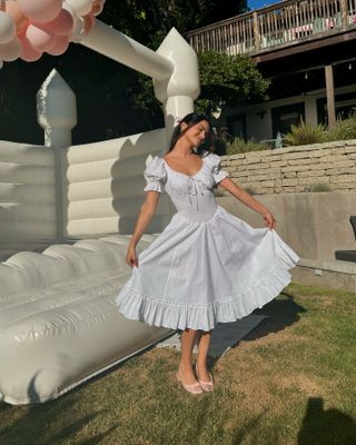 Camila Mendes wears a white midi dress with a pink bow in her hair