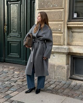 Danish art director Clara Dyrhauge poses on a cobblestone sidewalk wearing an oversize gray belted coat, a brown suede bag, straight-leg jeans, and black kitten-heel boots.