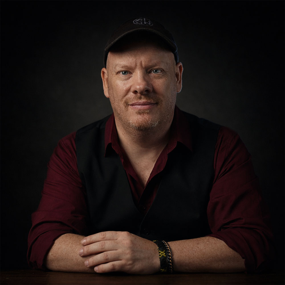 A man in a dark vest and maroon shirt sits at a table with his hands clasped, set against a shadowy background