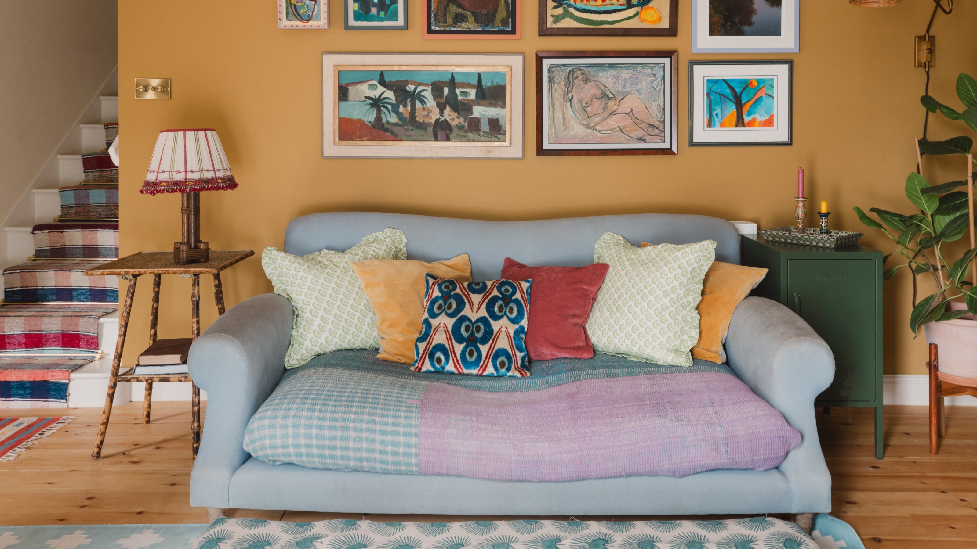 An ochre yellow-painted living room with a two-seater sofa with colourful cushions and a gallery wall above it