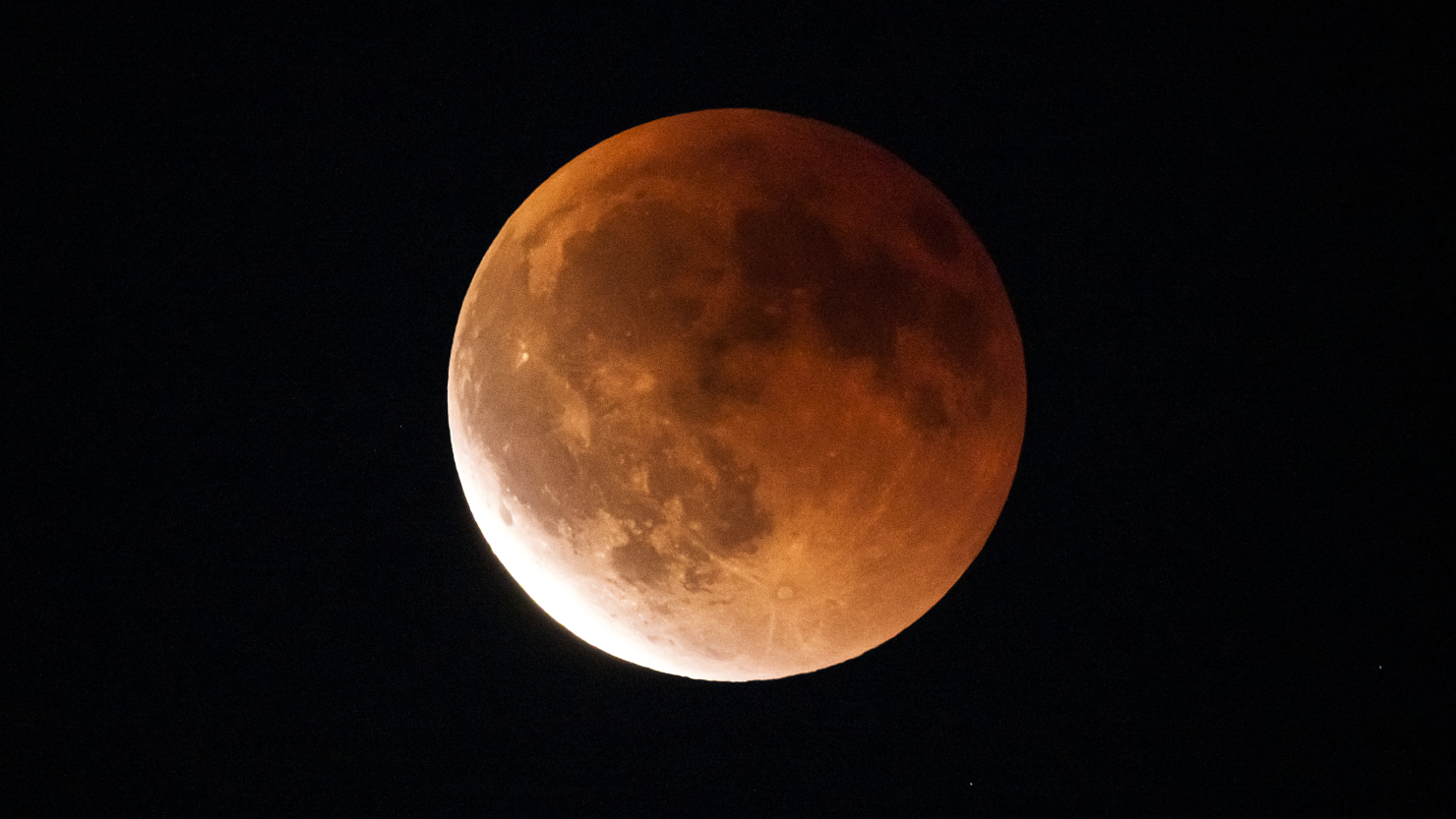 A yellow-orange eclipsed moon is pictured against a dark sky, with dark features known as lunar maria marking its surface along with the shapes of bright impact craters. The lower left part of its disk is brighter than the rest of its surface.