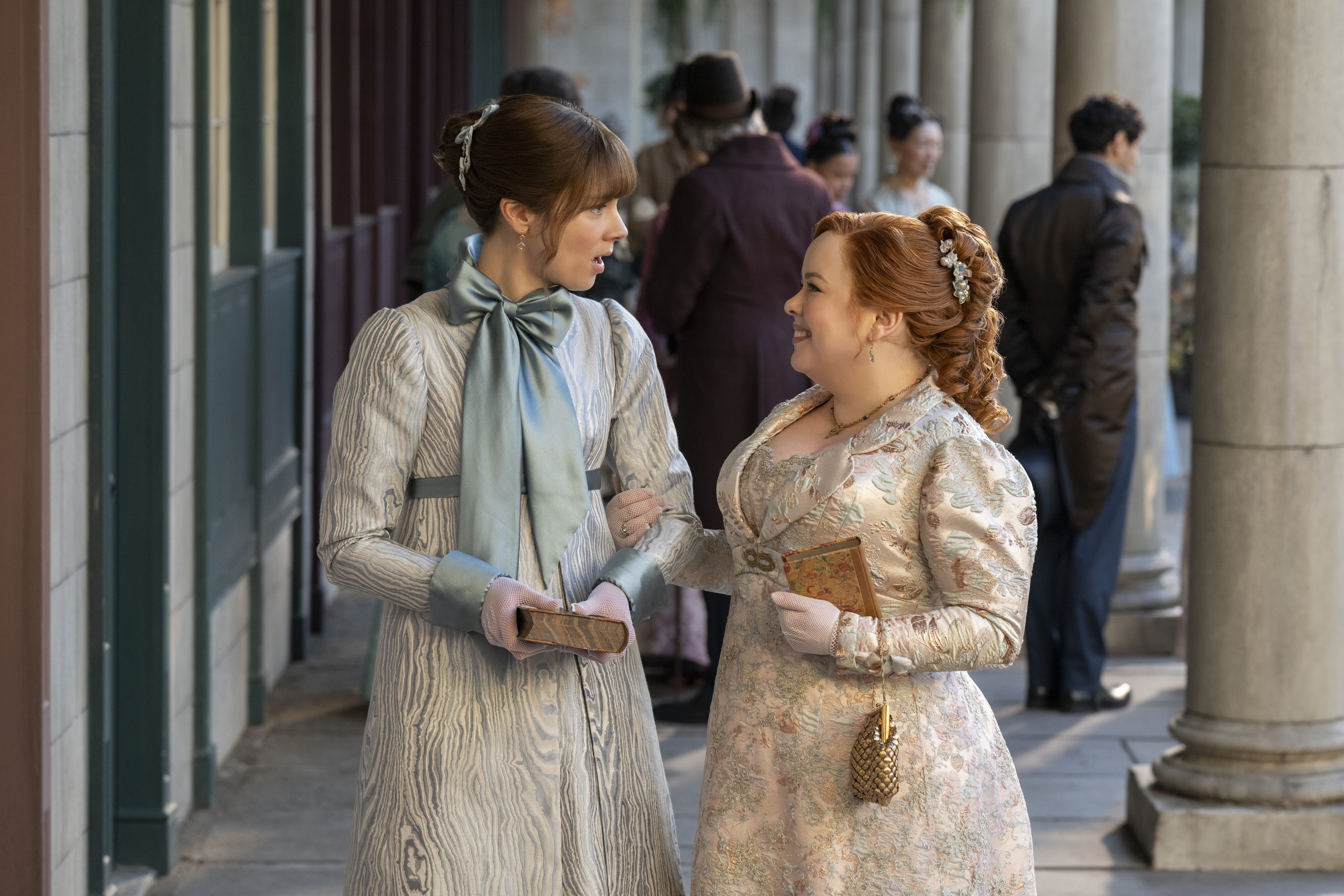 Claudia Jessie as Eloise Bridgerton wearing a blue dress with a bow around the neck and Nicola Coughlan as Penelope Featherington wearing a silver gown walking arm and arm through town in bridgerton season 4