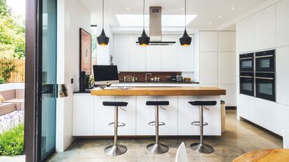 a large white kitchen extension with contemporary feel, wooden breakfast bar and wooden stools by keith collie, four ovens, black bar stools and black hanging lights – a good example of looking for how to add value to your home