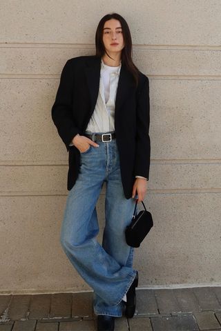 how to make a button-down shirt look more expensive is shown in a photo of a woman standing outside against a wall wearing a black blazer over a gray and white striped button-down shirt that's layered over a white t-shirt both of which are tucked into a pair of baggy jeans that are styled with a black belt, black loafers, and a black barrel bag