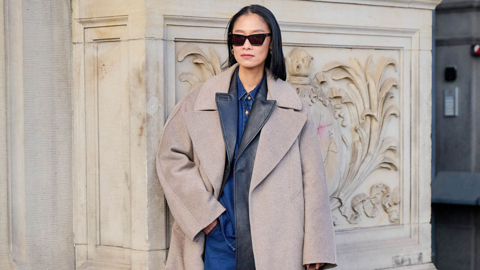 A woman is pictured wearing a blue denim set with a black leather blazer layered over it. Her outfit also includes an oversized beige coat with a large handbag and black boots. 