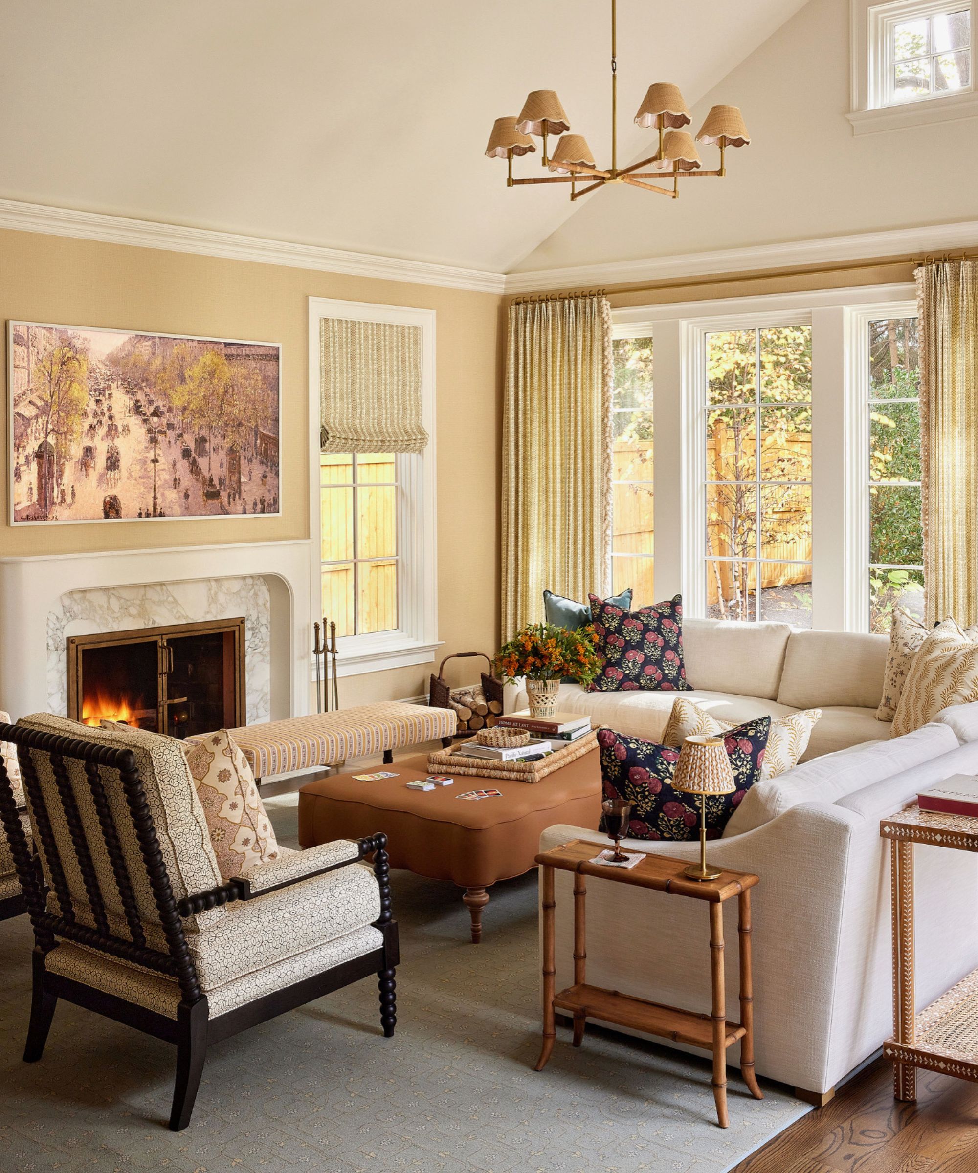 Living room with warm beige walls, large French doors with floral curtains, cream sectional sofa, brown leather ottoman, bamboo side table beside the sofa, and a bobbin armchair beside a large marble fireplace with a hidden TV above it