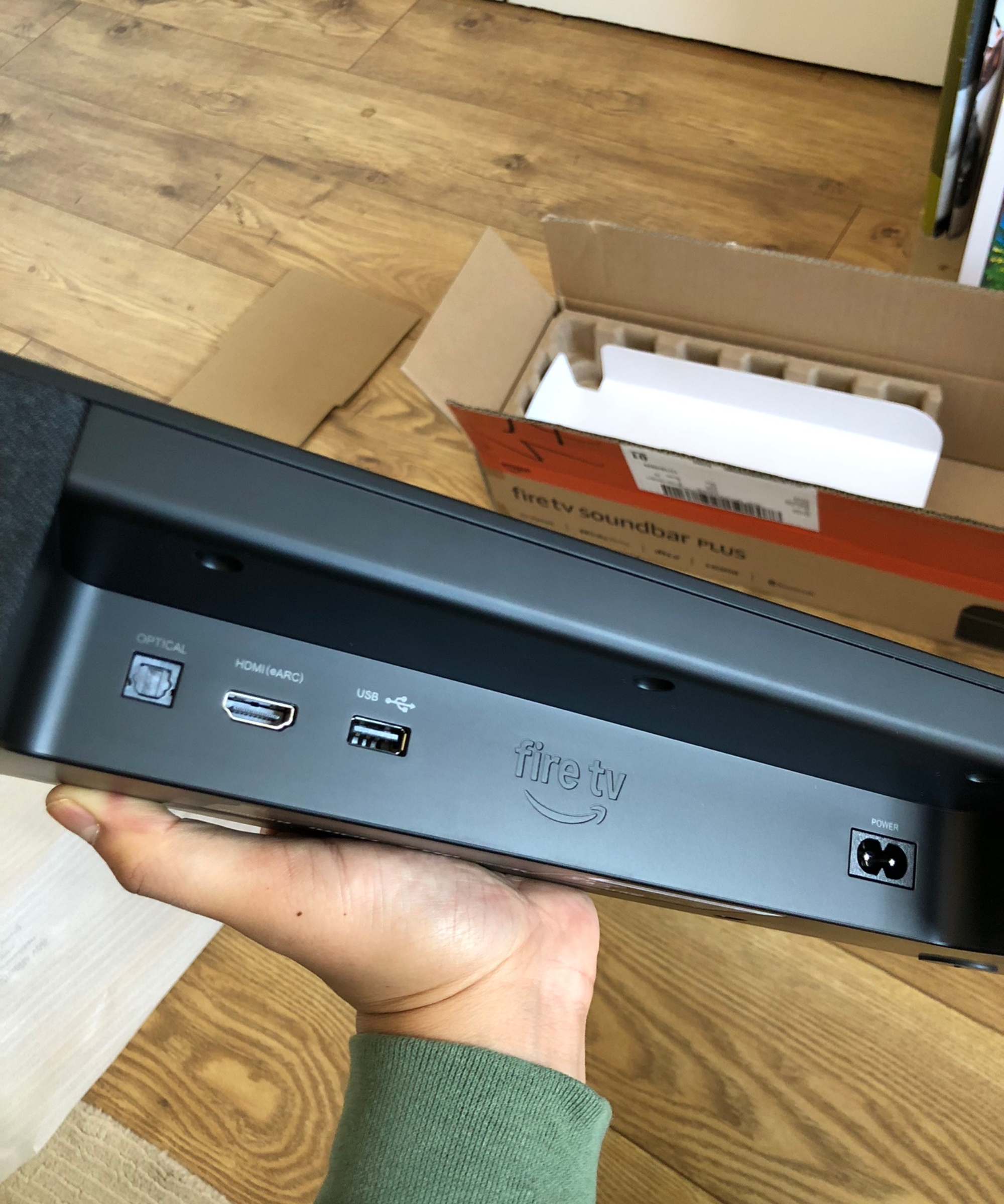 Hand holding the Fire TV Soundbar Plus showing the optical, HDMI, USB and power ports, with wooden floor in background in Dan Fauzi's home.