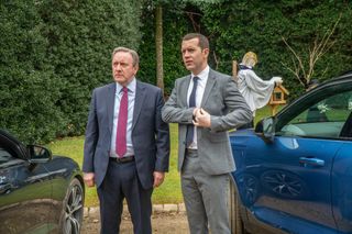 DCI John Barnaby (Neil Dudgeon) and DS Jamie Winter (Nick Hendrix) stand between their cars, a tableau of scarecrows behind them on the grass including one styled as a vicar. DS Winter is reaching into his inside jacket pocket.