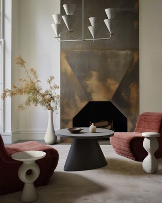 A white living room with a large muted metal fireplace that extends from floor to ceiling. There are two terracotta red velvet accent chairs, two white geometric side tables, and a black geometric coffee table on top of a white rug.