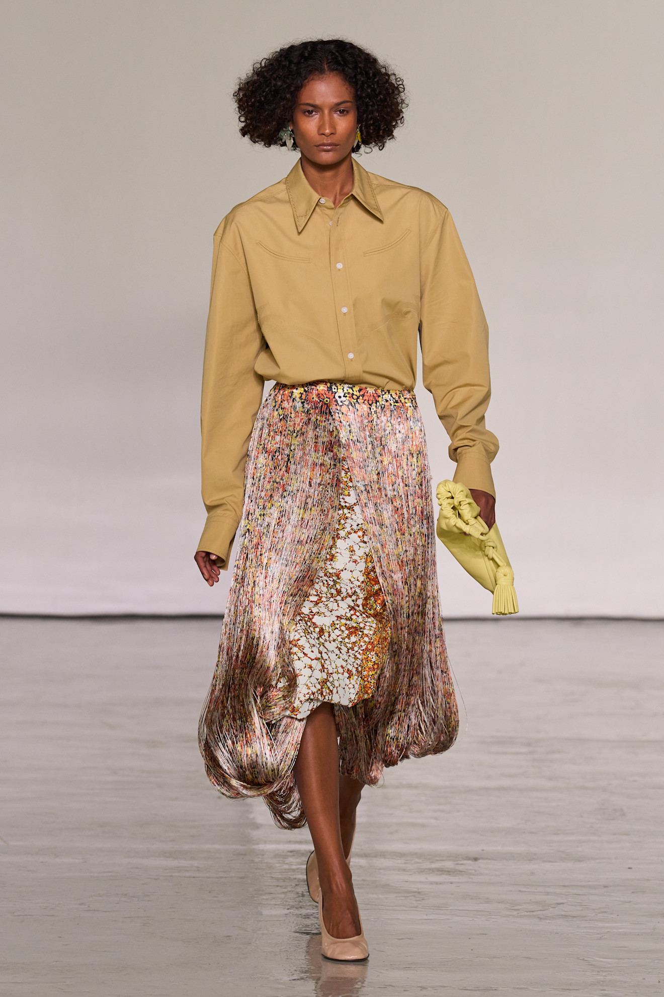A model walks the runway at Bottega Veneta's spring/summer 2027 Milan Fashion Week show wearing a mustard-colored button-down shirt with a multicolored fringe skirt.
