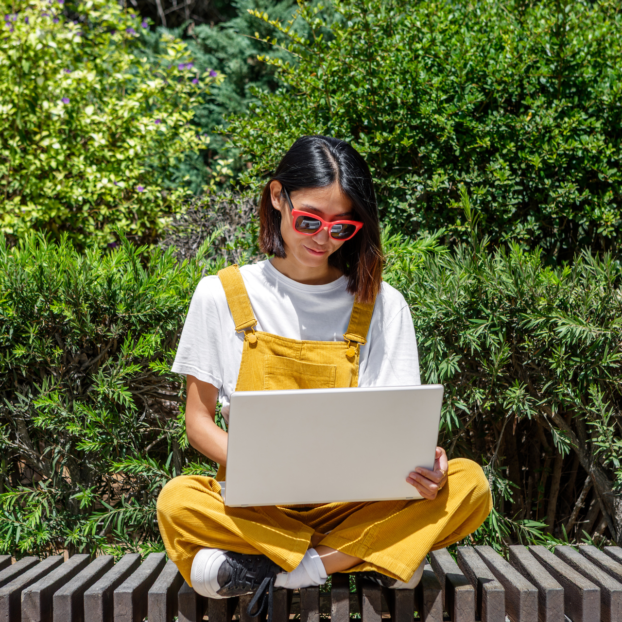 A woman sits cross-legged on a bench, wearing yellow overalls and sunglasses, while focused on her laptop. The setting is a lush green garden, indicating a warm, sunny day.