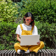 A woman sits cross-legged on a bench, wearing yellow overalls and sunglasses, while focused on her laptop. The setting is a lush green garden, indicating a warm, sunny day.