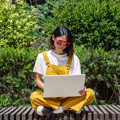 A woman sits cross-legged on a bench, wearing yellow overalls and sunglasses, while focused on her laptop. The setting is a lush green garden, indicating a warm, sunny day.