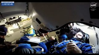 a man in a blue spacesuit sits scrunched inside a spacecraft. A pair of black-clad individuals assist him.