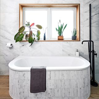 a curved bathtub under a window in a bathroom, clad in marble pencil tiles