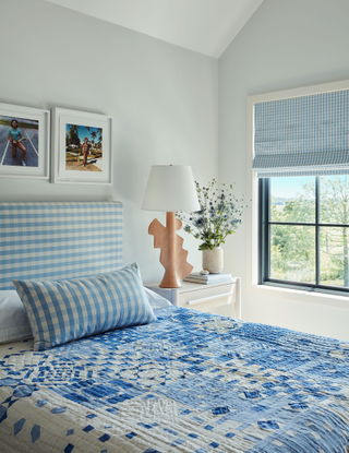 A white bedroom with light blue details. The bed is adorned with a blue and white quilt and a headboard and throw pillow in a coordinating, lighter blue gingham pattern. There are photos hung above the bed and a white beside table with a clay, geometric table lamp.