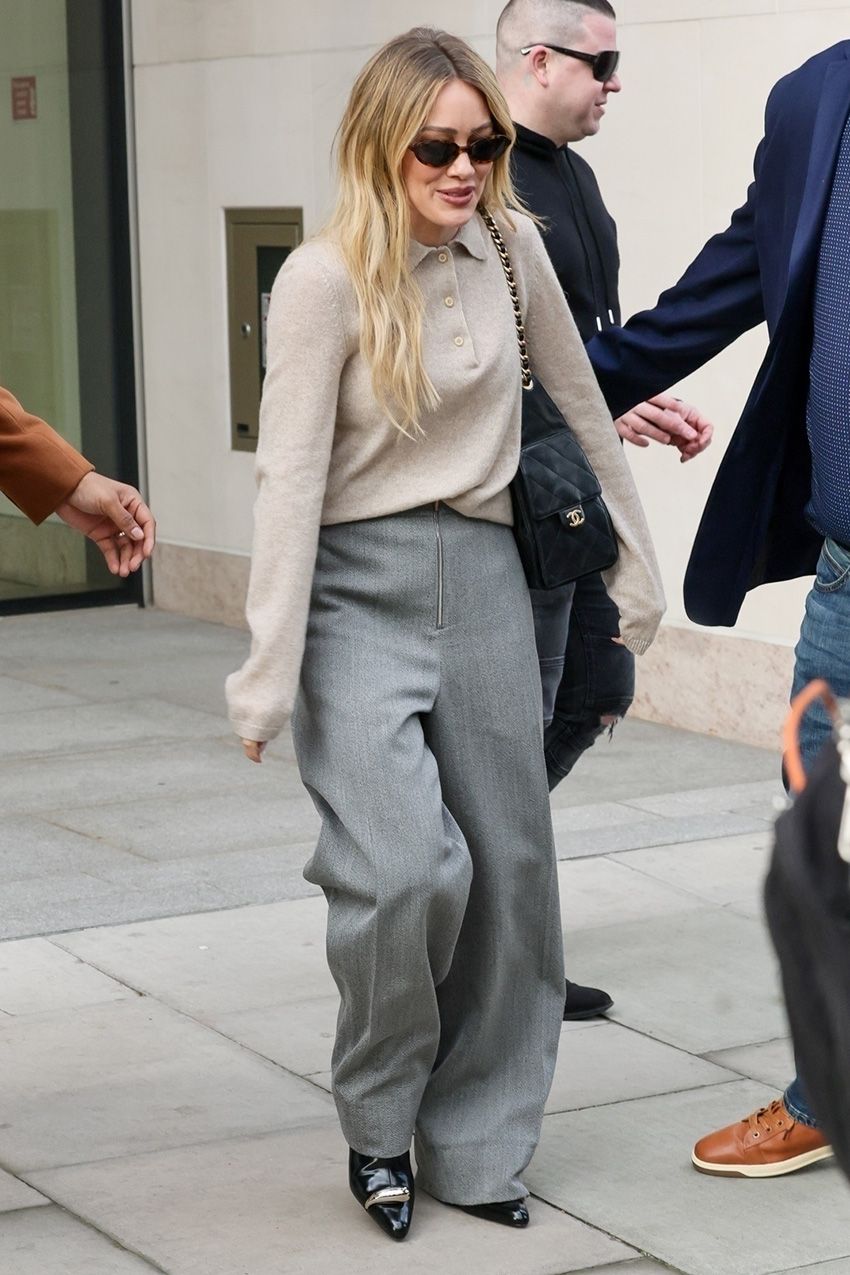 Hilary Duff wearing an oatmeal polo sweater, gray trousers, a Chanel 25 bag, and black pointed-toe shoes in London