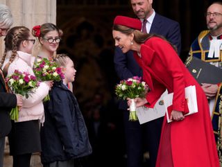 Kate Middleton wearing a red coat and hat leaning forward and taking flowers from a little girl at Westminster Abbey