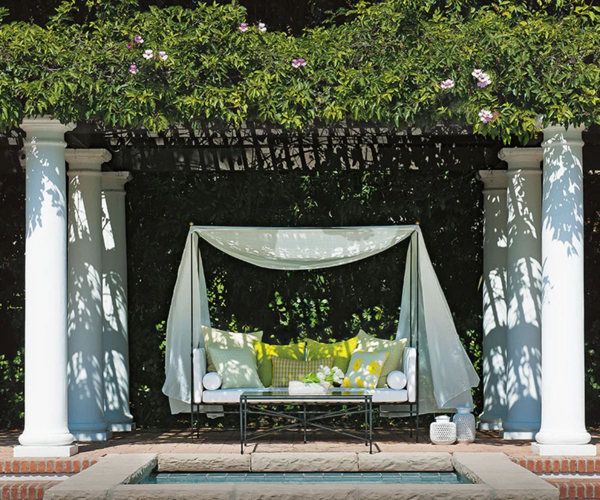 outdoor garden poolside patio area with a covered canopy lounger sofa surrounded by greenery and pillars