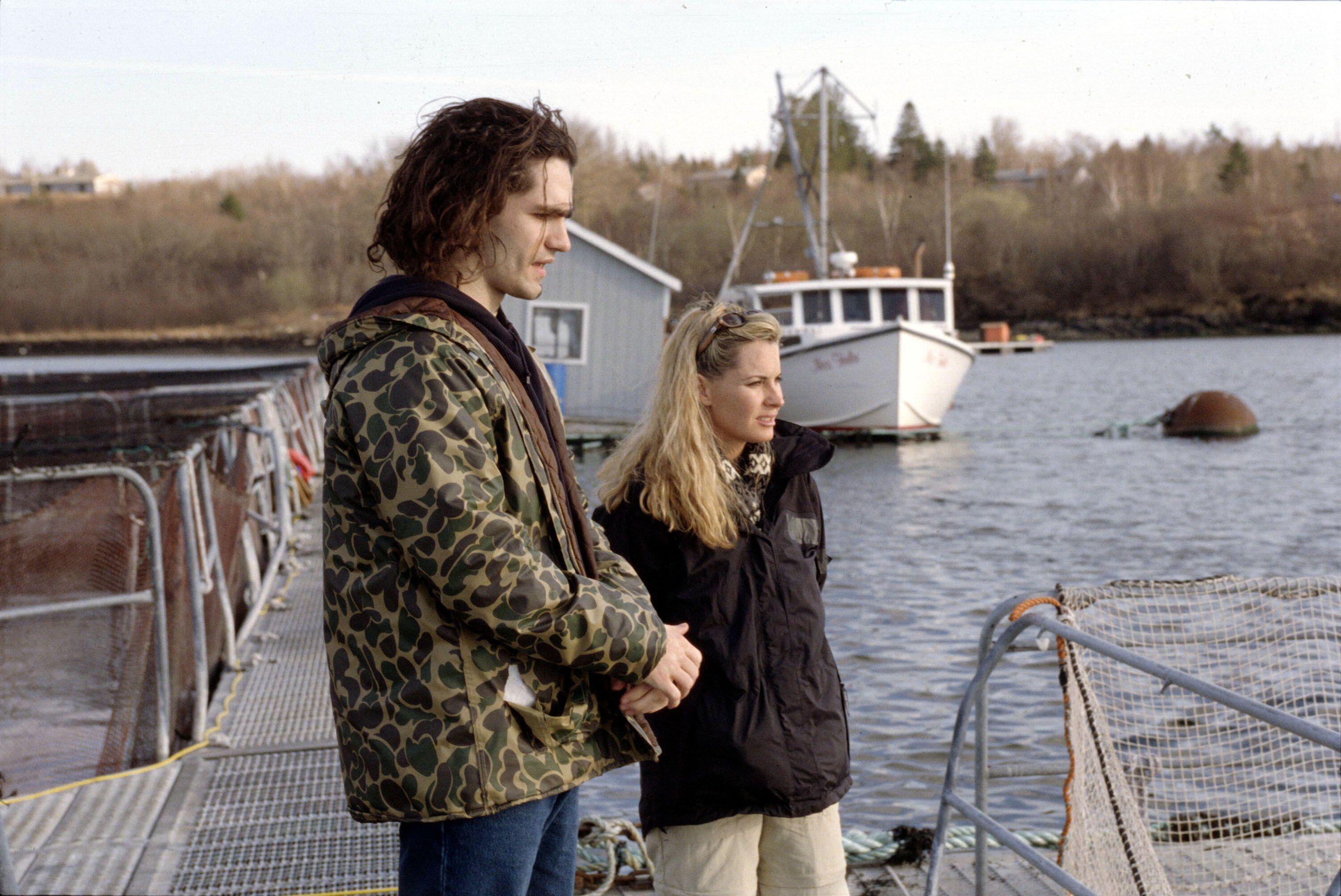 a man in a camo jacket and a a woman in a black jacket stand by a pier in 'Murder In Small Town X'