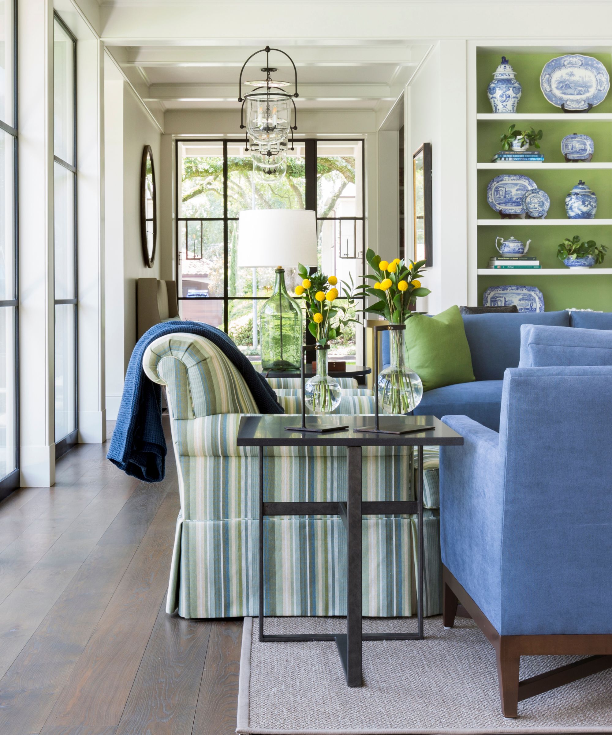 neutral traditional living area with large glass windows, a green and blue striped couch with blue arm chairs and bookshelves painted apple green with blue and white vintage crockery