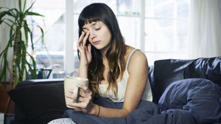 Woman sat up, rubbing her eyes and looking tired, with a mug in her hand