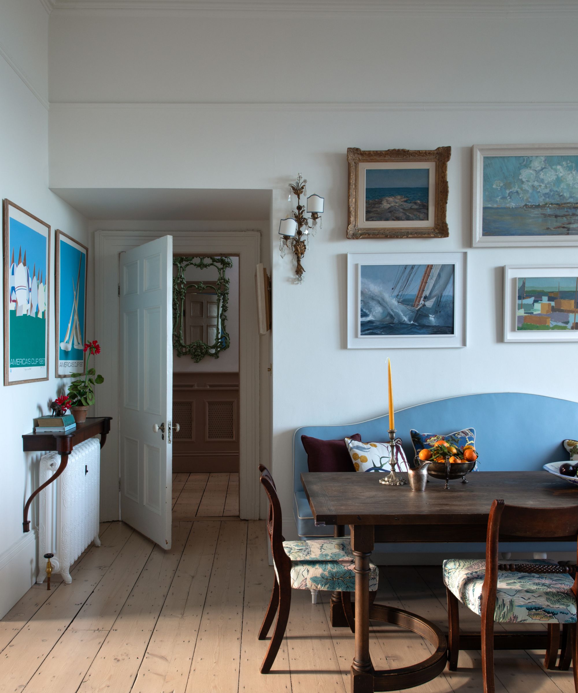 Dining room with wooden floorboards, gallery wall of seascapes above the dining table, blue banquette bench, mahogany table and chairs and antique candlesticks and bowl