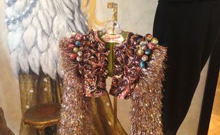 Close up image of a flambouyant shrug cardigan with long sparkle fibre sleeves, gold stool, black trouser hung up, wall angel wings, with gold cloak painted art work on the back wall