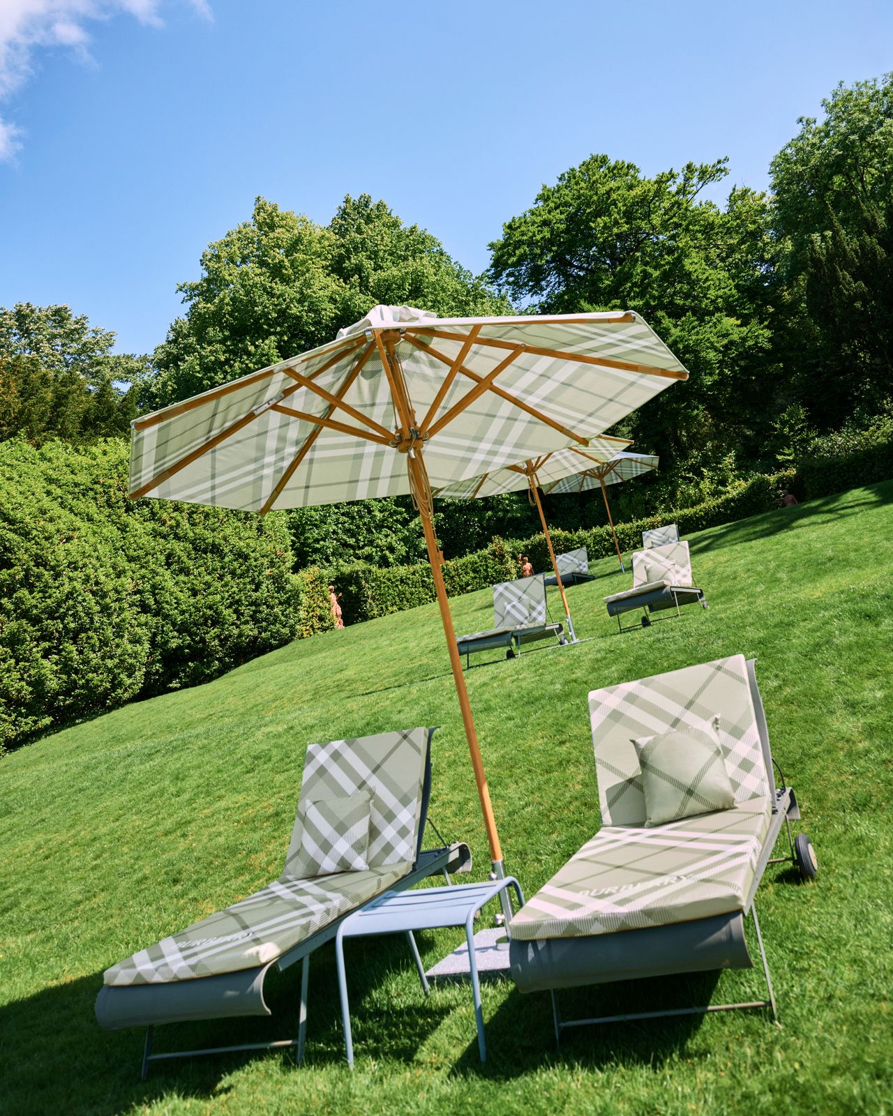 A green lawn with three rows of sun loungers covered in a pale green Burberry plaid with wooden parasols, also patterned in plaid