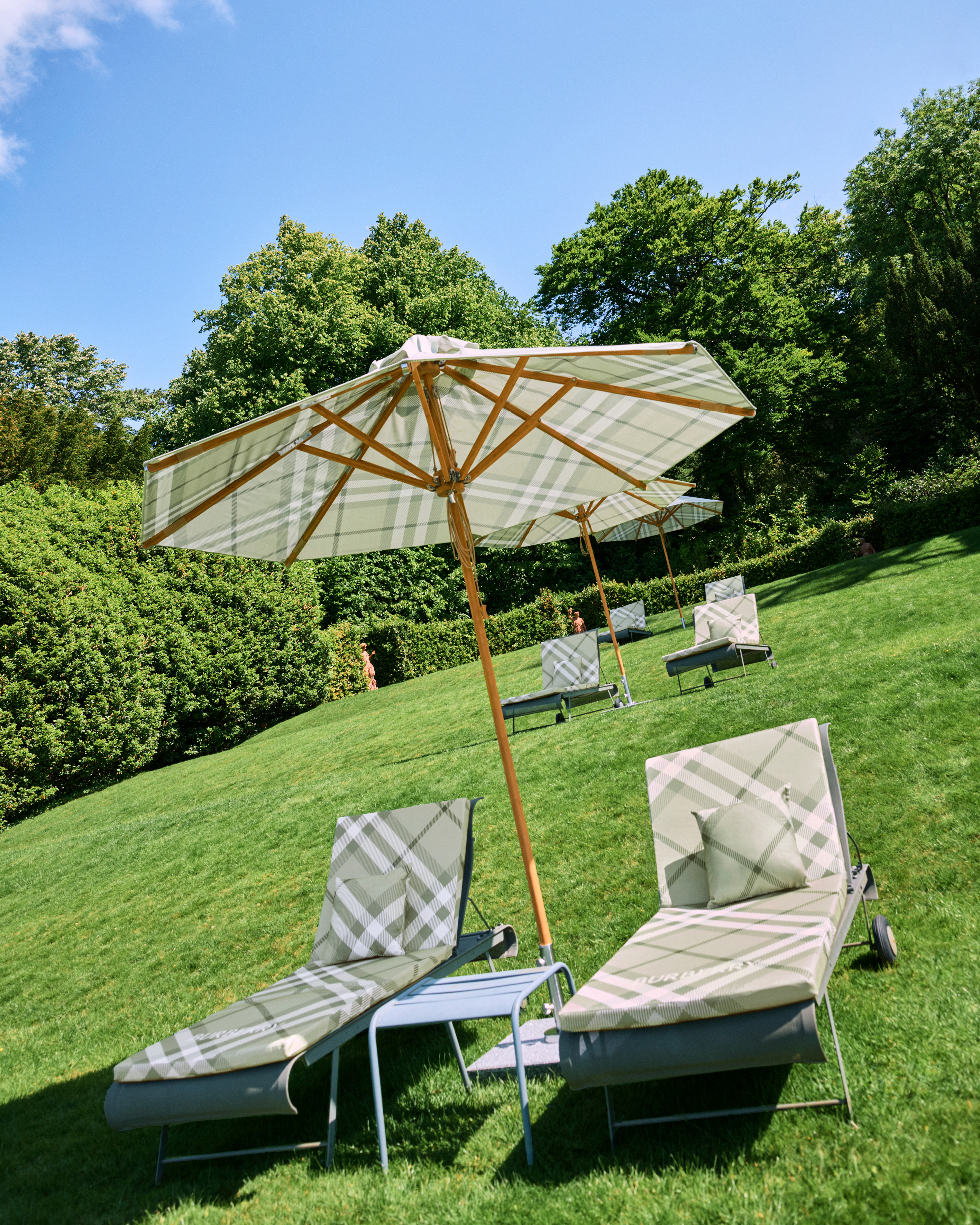 A green lawn with three rows of sun loungers covered in a pale green Burberry plaid with wooden parasols, also patterned in plaid