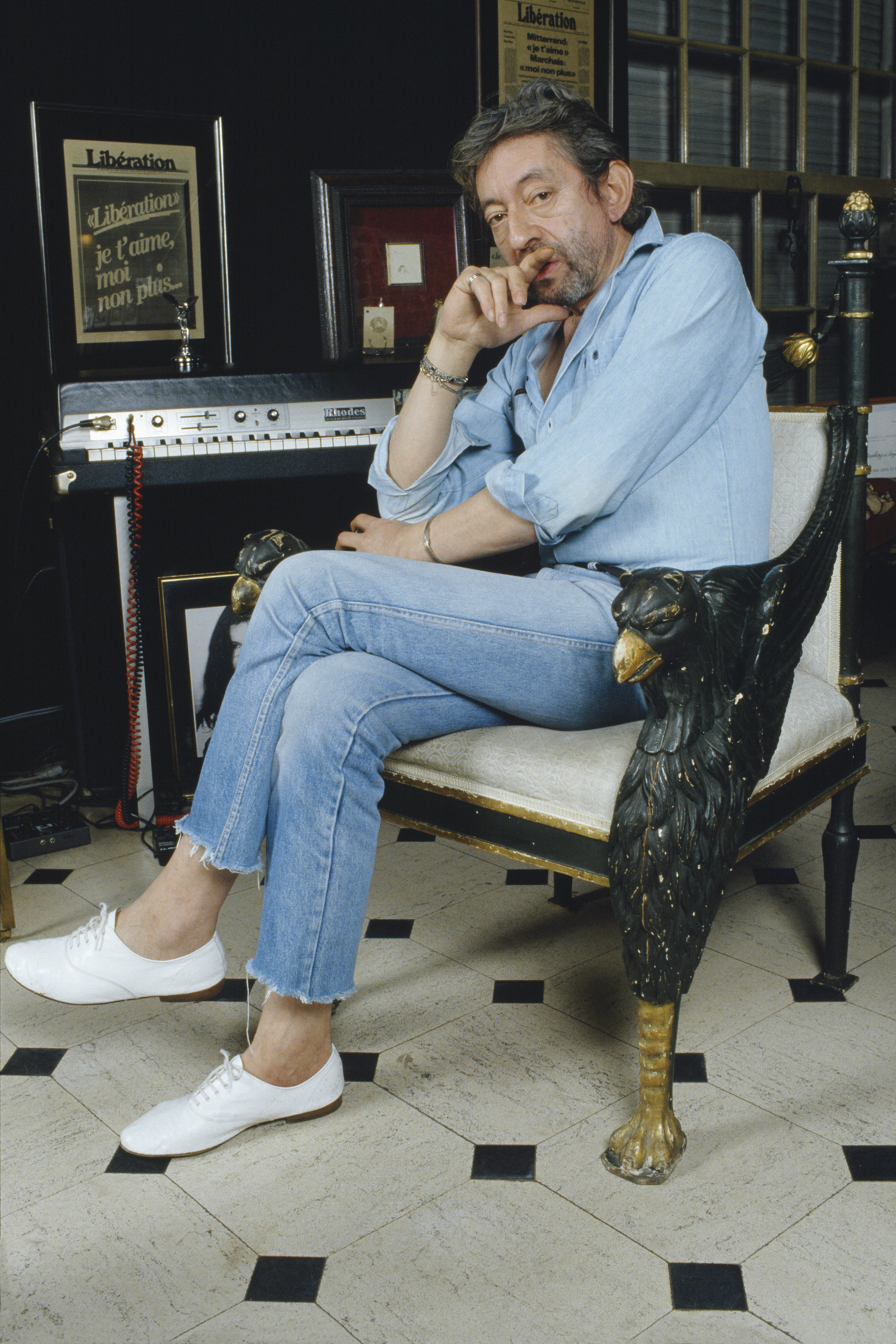 Serge Gainsbourg sitting on an ornate chair wearing a denim shirt with jeans and white lace-up oxford shoes
