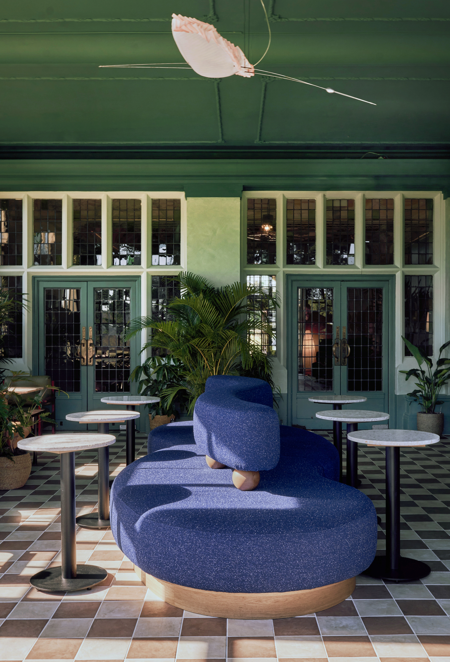 double-sided blue sofa with wavy backrest in orangery with checkered floors, bar tables, and potted plants, with the ceiling and walls painted green