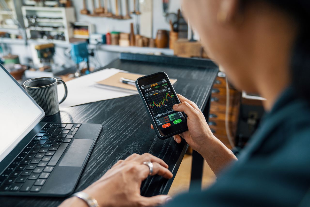 Man looks at stocks and shares on mobile phone as he sits at desk beside laptop.