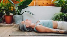 Woman lying down on her back on yoga mat at home surrounded by plants, doing pelvic tilt exercise