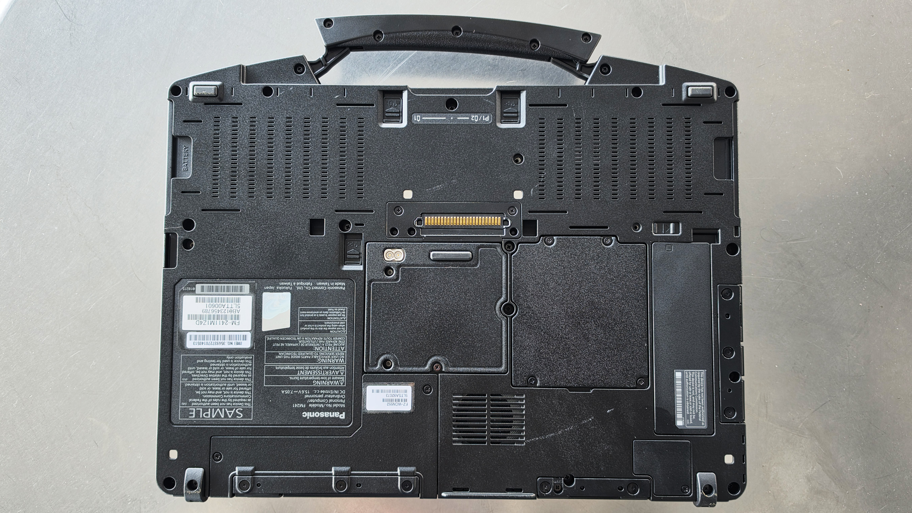 The image shows the underside of a rugged black laptop, with visible screws, ventilation grills, and labeled compartments on a gray surface.