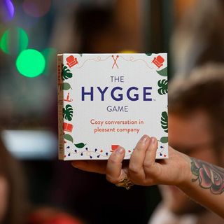 A person's hand holding a white card game box titled, "The Hygee Game" 