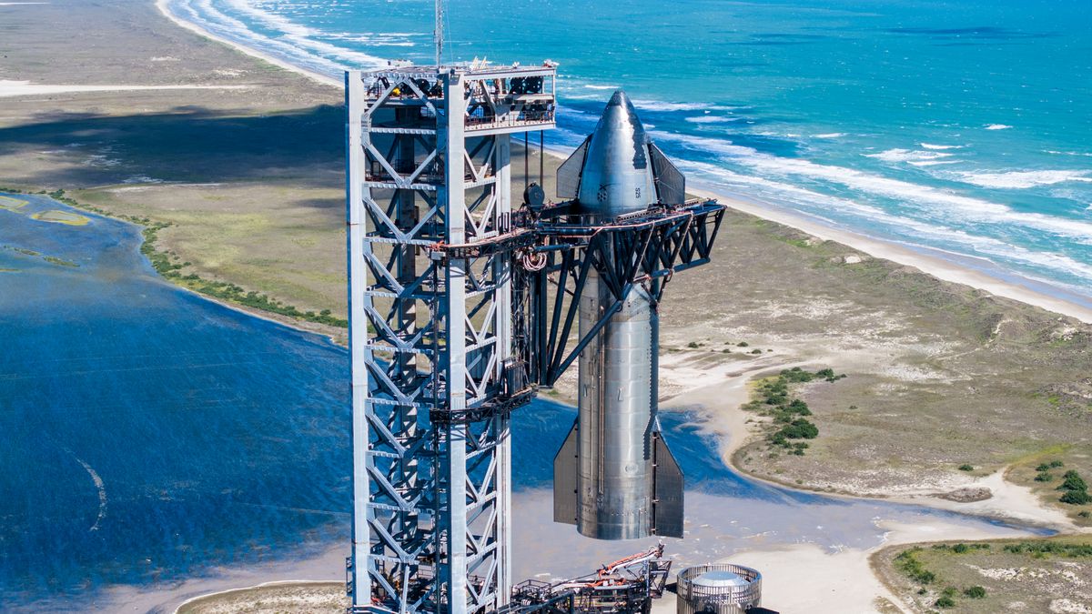 a silver rocket is held against its gray launch tower, with the turquoise waters of the Gulf of Mexico in the background.