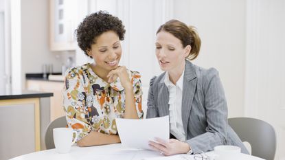 A financial adviser and her client look over paperwork at a table in an office.
