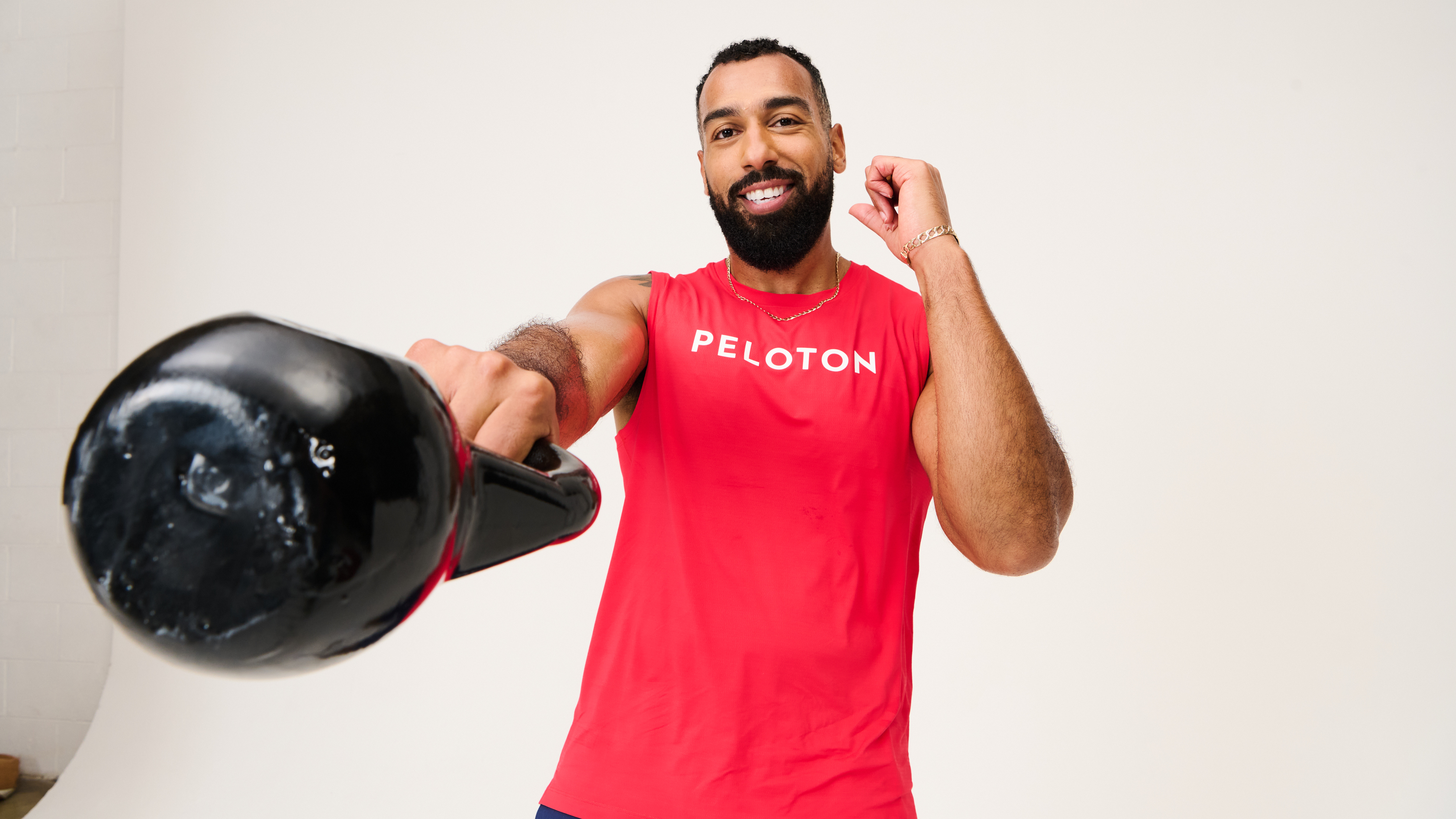 Man performing single-arm kettlebell swing wearing red vest with Peloton written in white