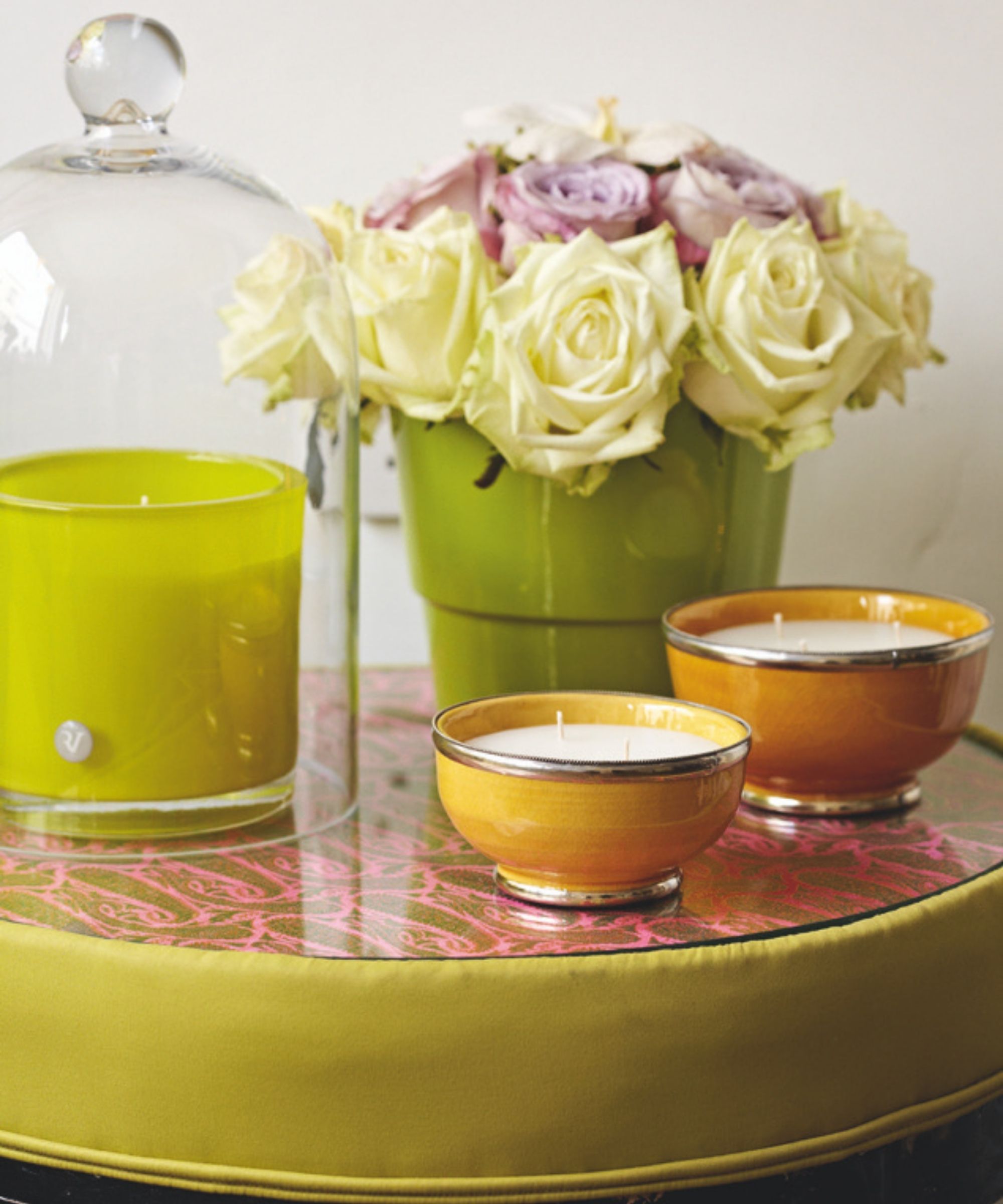 Pink and white roses in a green pot on a green table with a pink and green top, with two small yellow candles with white wax, and a green candle under a glass cloche. The background is white.