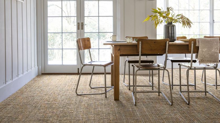 A light-filled dining room featuring a rustic wooden table and vintage metal chairs on a textured woven carpet. Large French doors flood the space with natural light, highlighting the room’s relaxed, natural style.