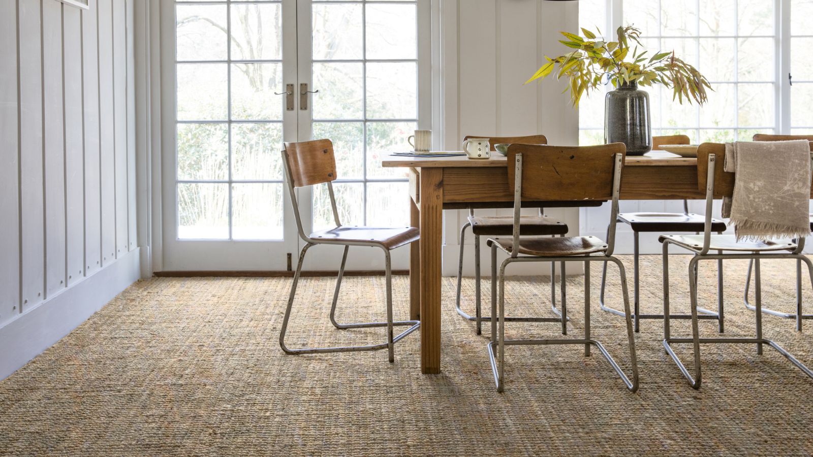 A light-filled dining room featuring a rustic wooden table and vintage metal chairs on a textured woven carpet. Large French doors flood the space with natural light, highlighting the room’s relaxed, natural style.