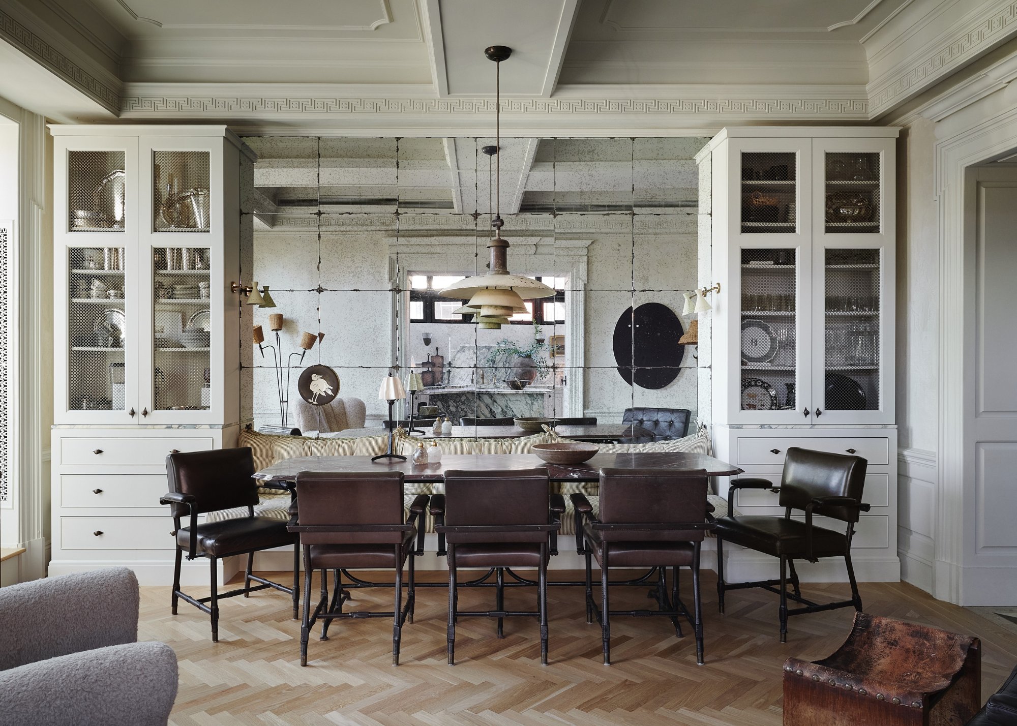 A dining space with mirrored walls flanked either side with glass-fronted shelving housing crockery and glassware and brown leather seats sit around one side of the table with in-built seating along the back wall on top of herringbone flooring