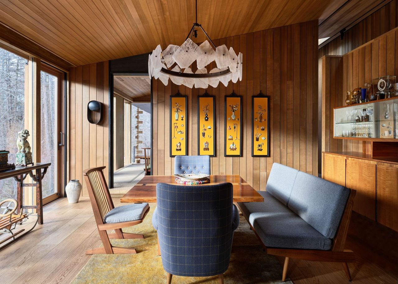 A Japanese dining room with wood paneled walls and sloped ceiling, a white chandelier above the wood table, four pieces of yellow-hued artwork hang on the walls and various blue seats around the table