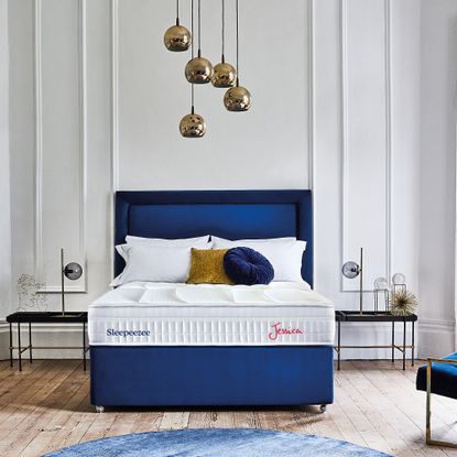 A navy blue upholstered bed holding the Jessica 1800 Pocket Gel Mattress. A light fixture with five brass globes hangs overhead and there is a white panelled wall behind the bed and a wooden floor