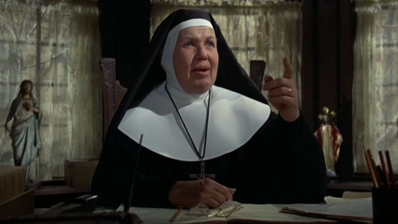 Kathleen Freeman gestures upward while sitting at a desk dressed as a nun in The Blues Brothers.