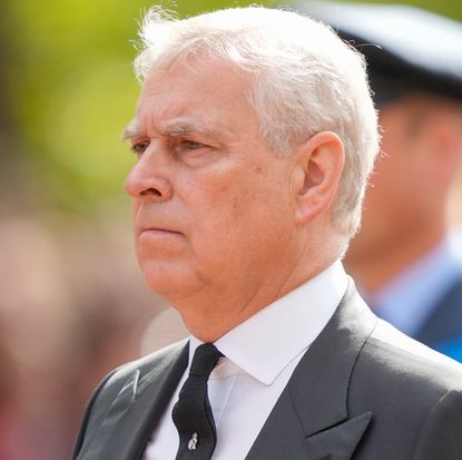 Prince Andrew wearing a gray suit and walking outside with a grim expression