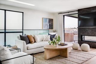 white apartment living room with view outside of outdoor kitchen, wooden oval coffee table, white couch and two armchairs, coir rug, glass, artwork, tv and modern fireplace