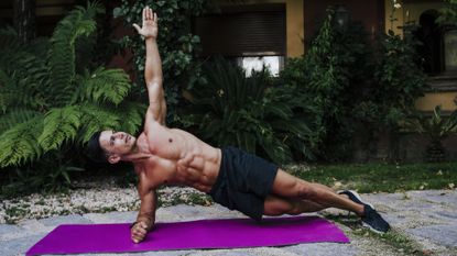 Shirtless male athlete practicing plank position on mat against plants in yard 